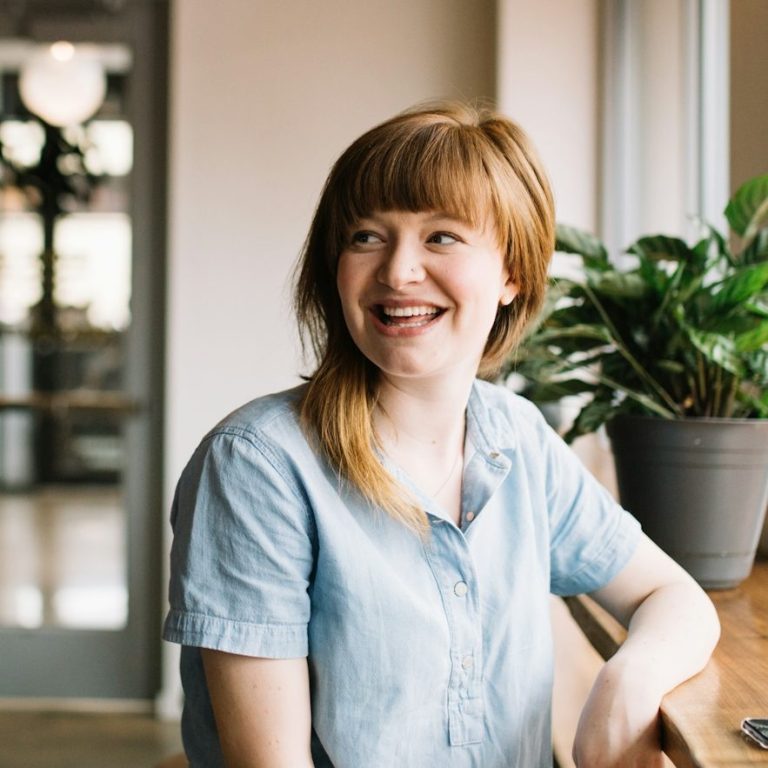 Femme souriante devant une fenêtre, avec une plante en pot à côté d'elle.