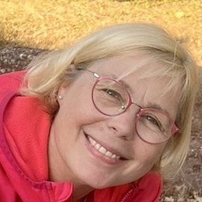 Femme souriante avec des lunettes roses, portant un pull rouge, posé sur un sol en terre.